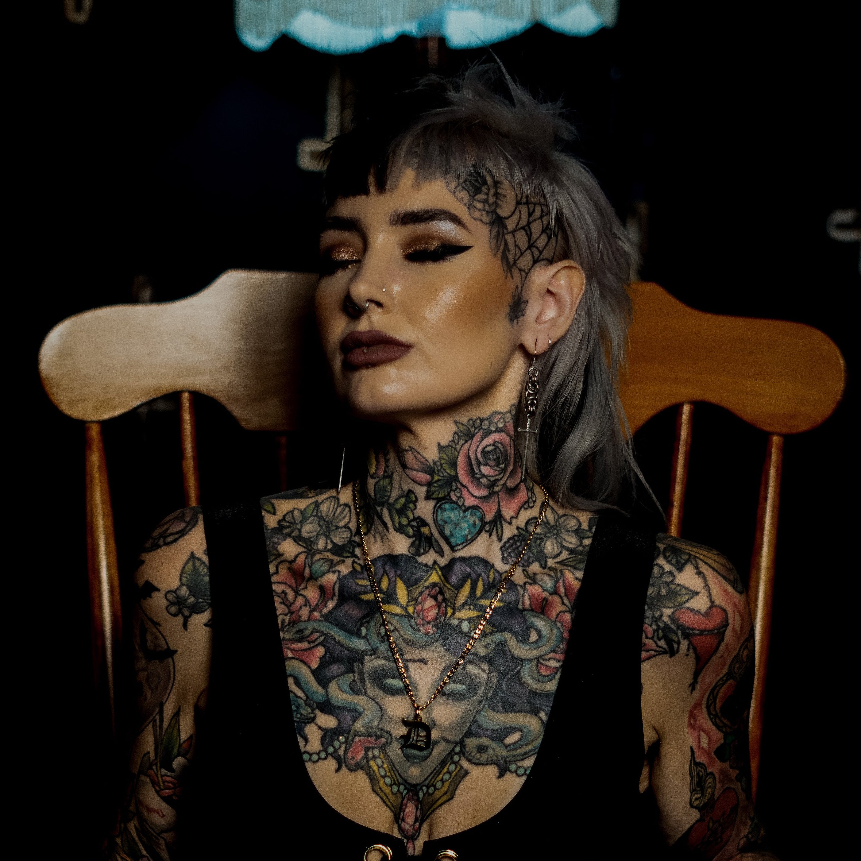Person with tattoos sitting in a chair under a lamp wearing a gold initial necklace