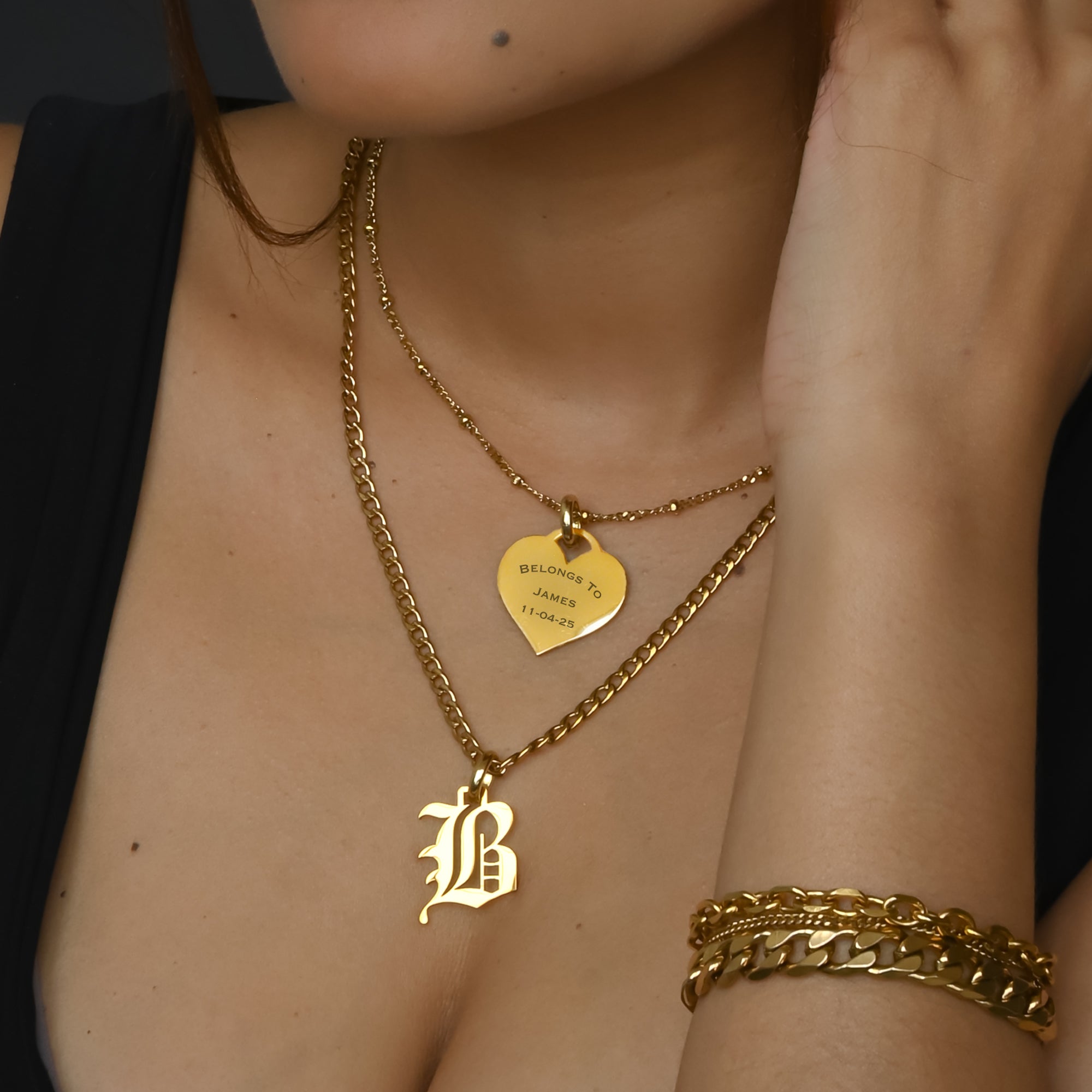 Gold necklace with a heart-shaped engraved pendant and 'B' pendant, worn by a person.
