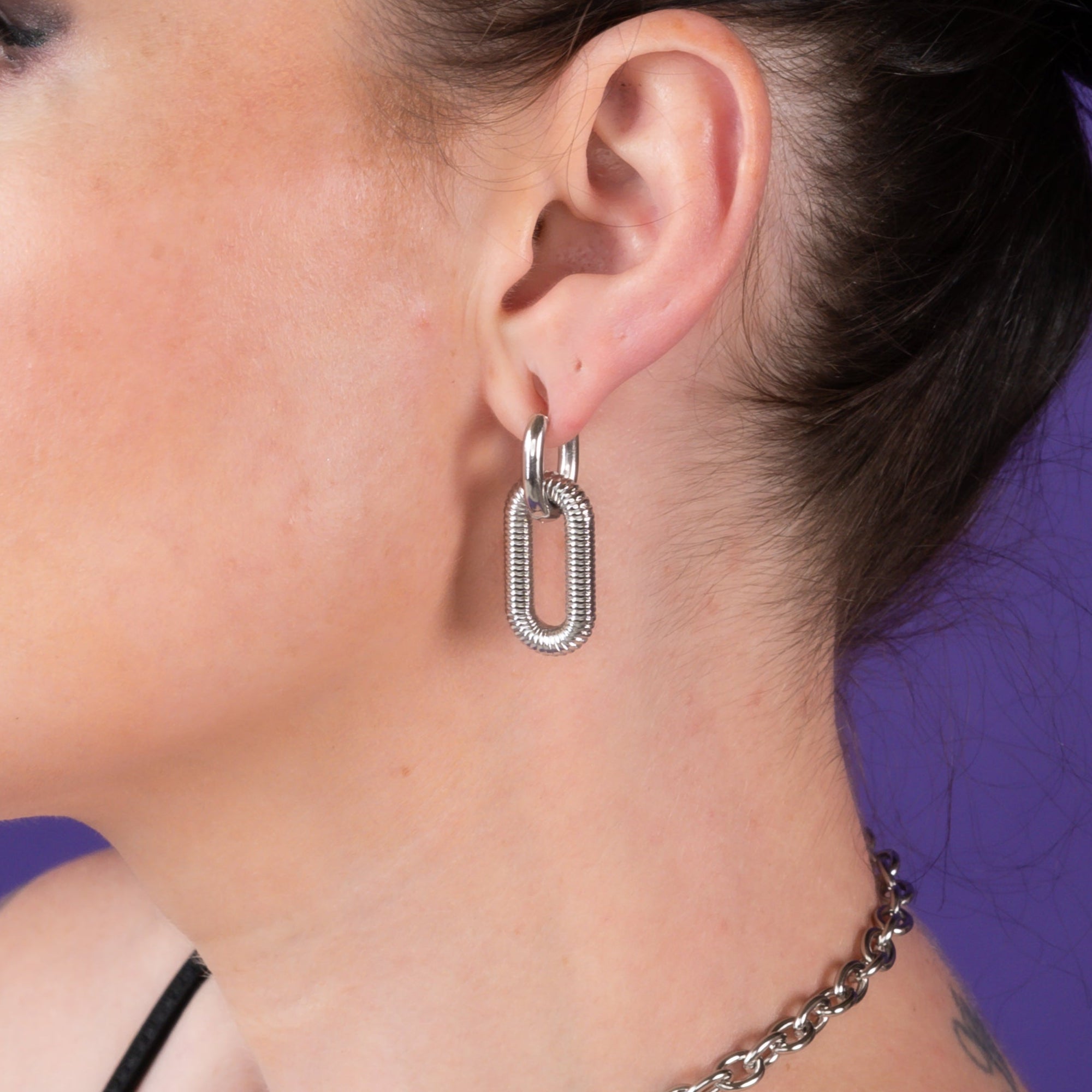 Woman with a bun hairstyle wearing silver chunky drop earrings and a choker against a purple background